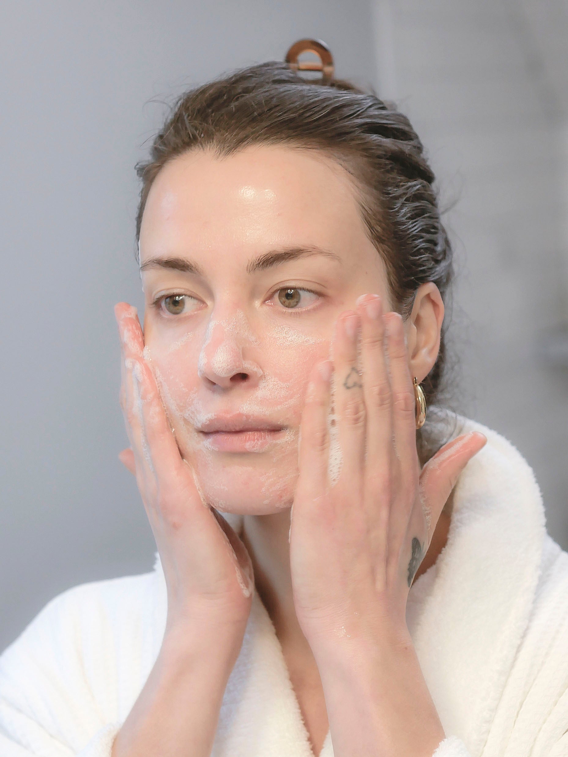 A woman with dark hair pulled back wearing a white fluffy bathrobe using day and night face cleansers.