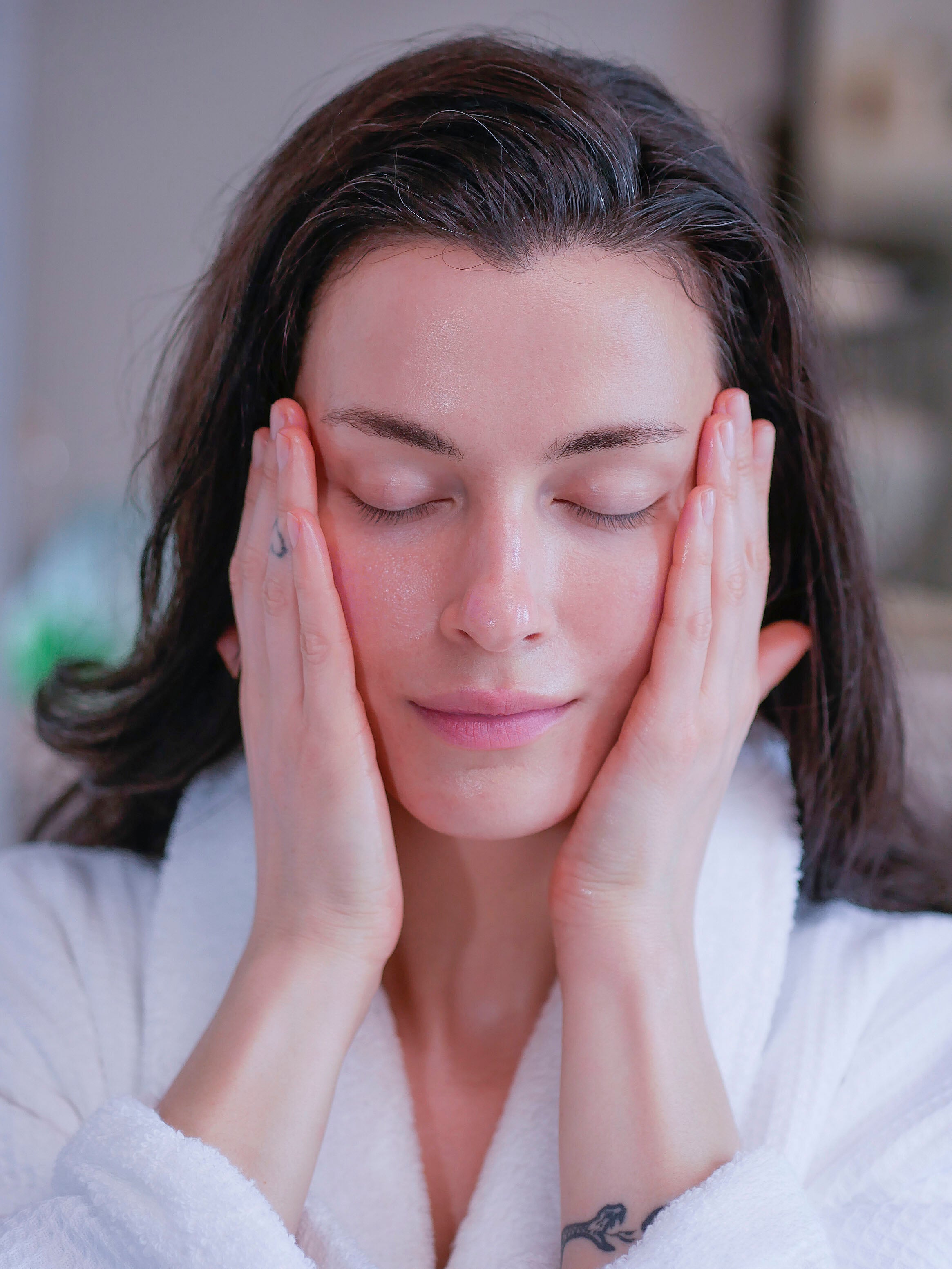 A woman with brown hair in a white fluffy bathrobe applying face serum for dry skin.
