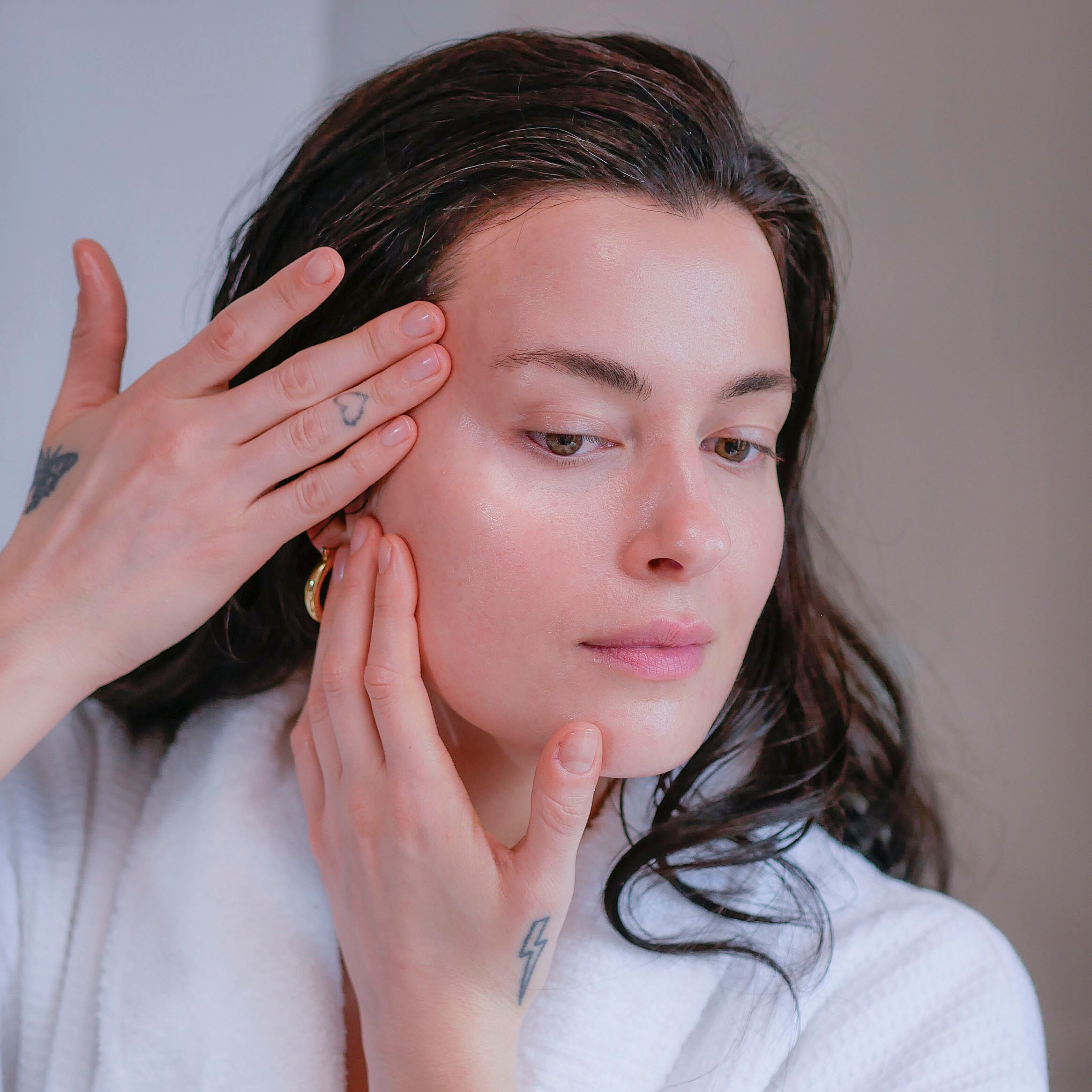 Woman with dark hair in a white fluffy bathrobe doing her natural skincare routine for her sensitive skin.