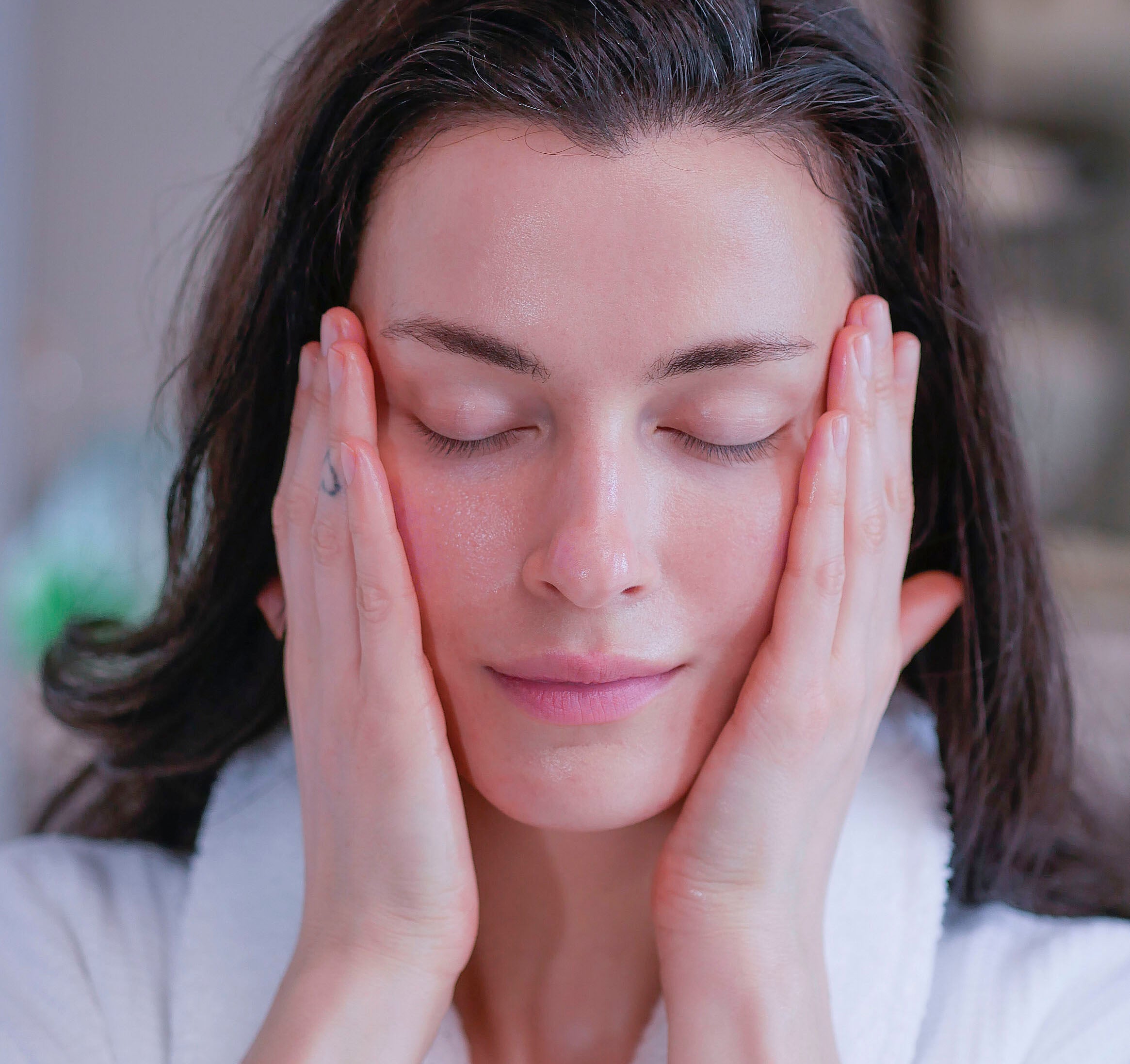 A woman with dark hair wearing a white fluffy bathrobe applying botanical skincare products from Cultivate Apothecary.