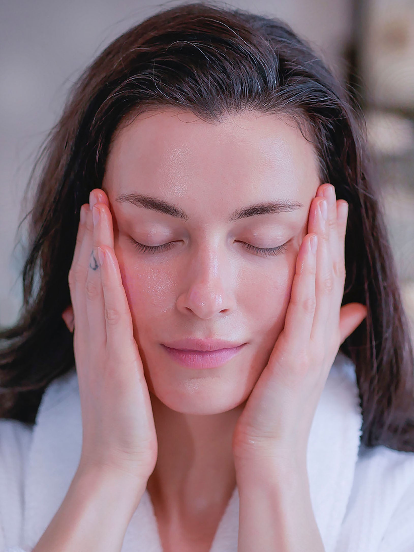 A woman with brown hair in a white fluffy bathrobe applying a botanical clean beauty facial toner for added hydration.
