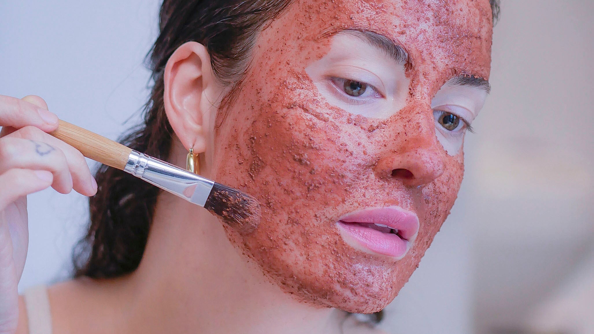 A woman applying a botanical and organic clay face mask using a face mask brush.