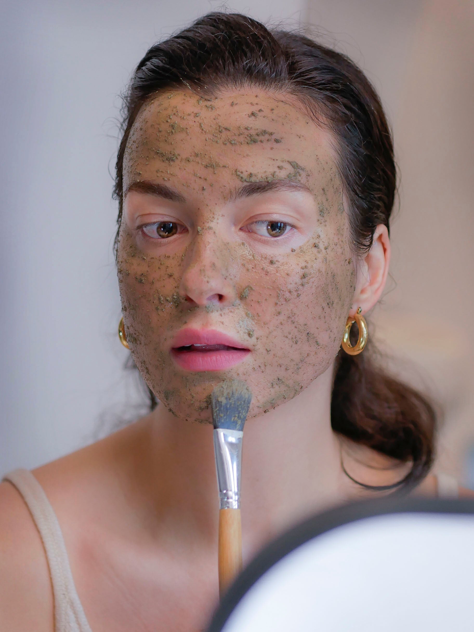 Woman with dark hair applying a healing botanical clay face mask for dry skin using a face mask brush.