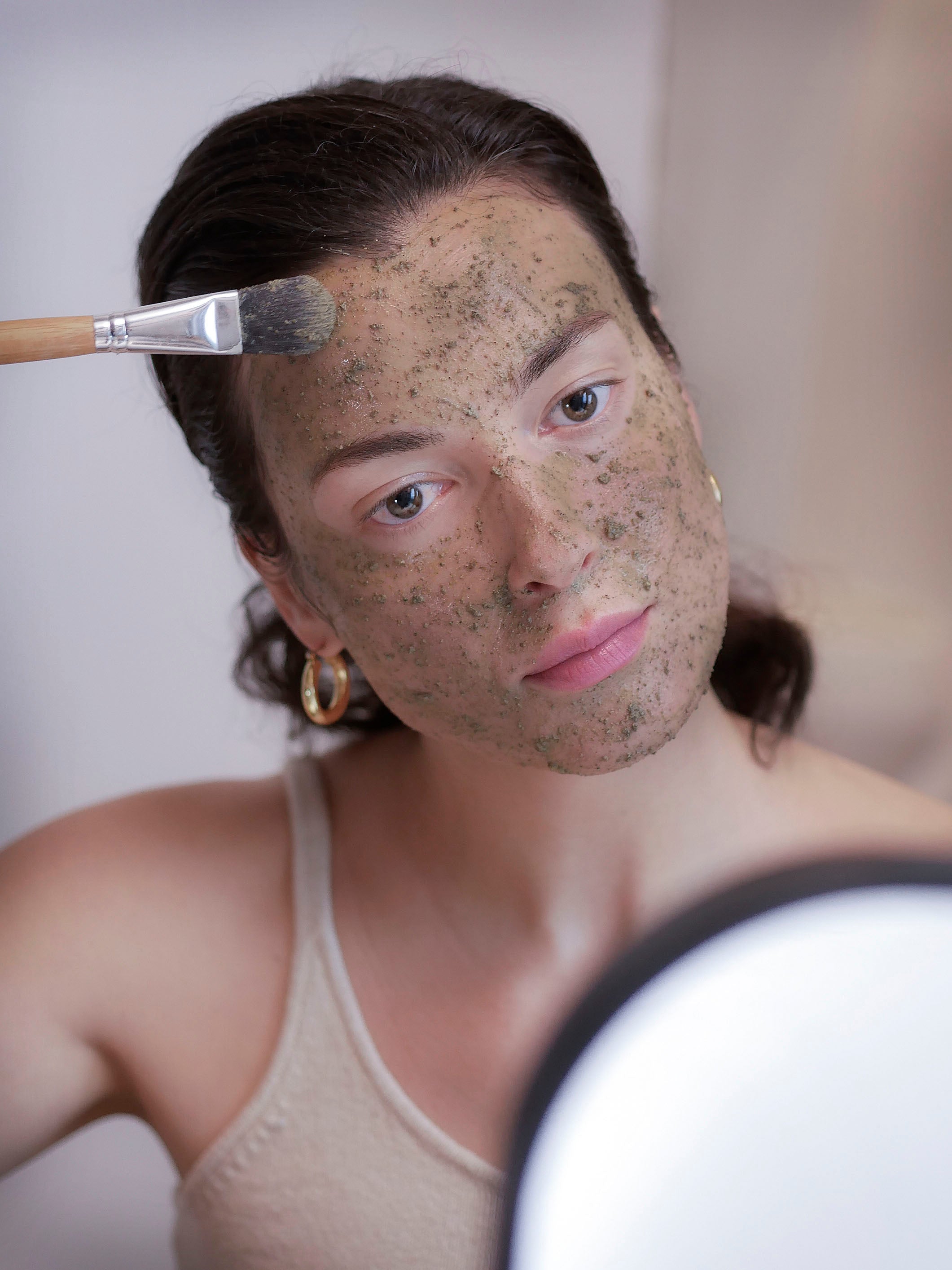 Woman with dark hair applying green clay face mask using a face mask brush.