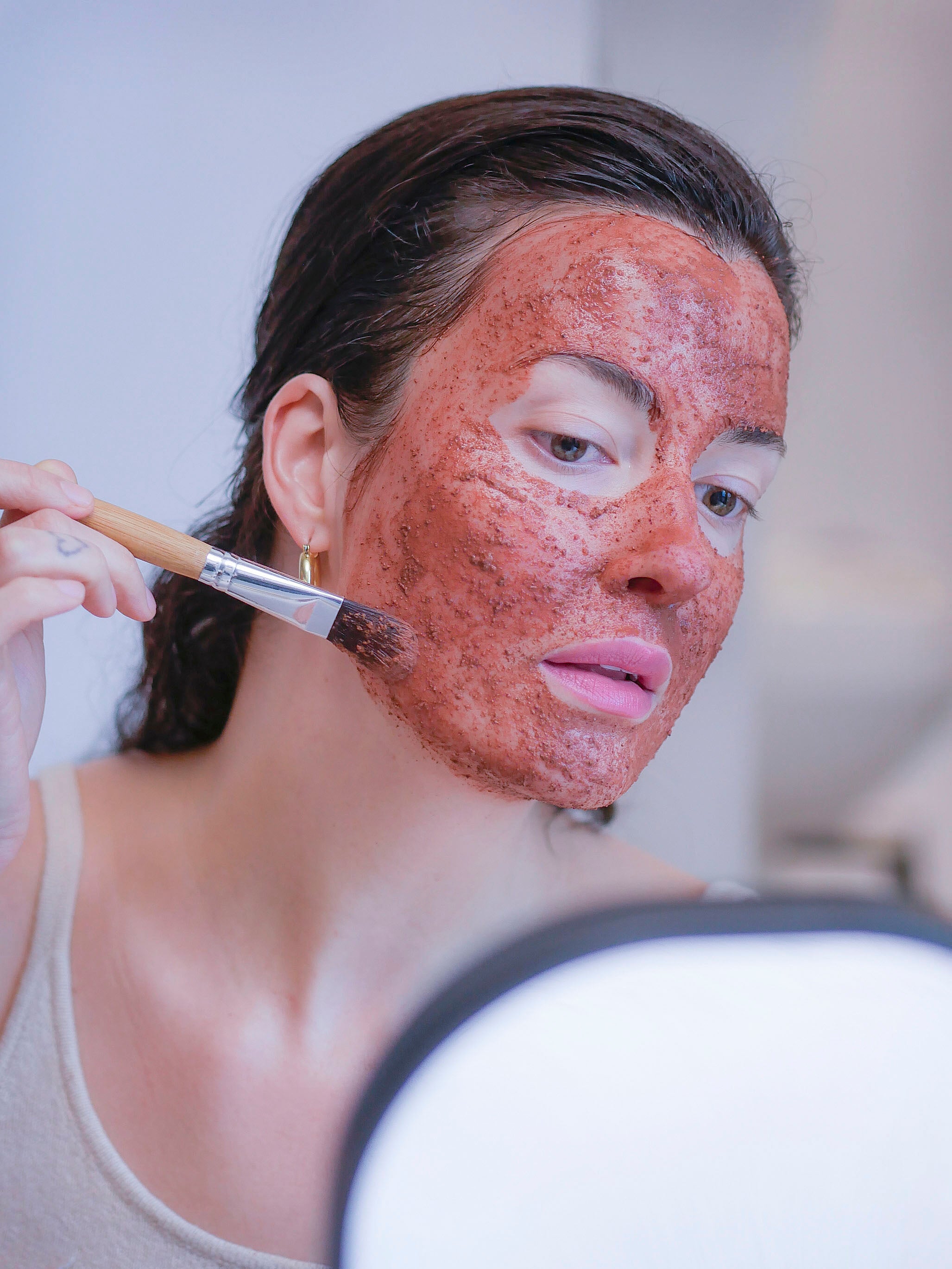Woman applying an exfoliating botanical pink clay mask using a face mask brush.