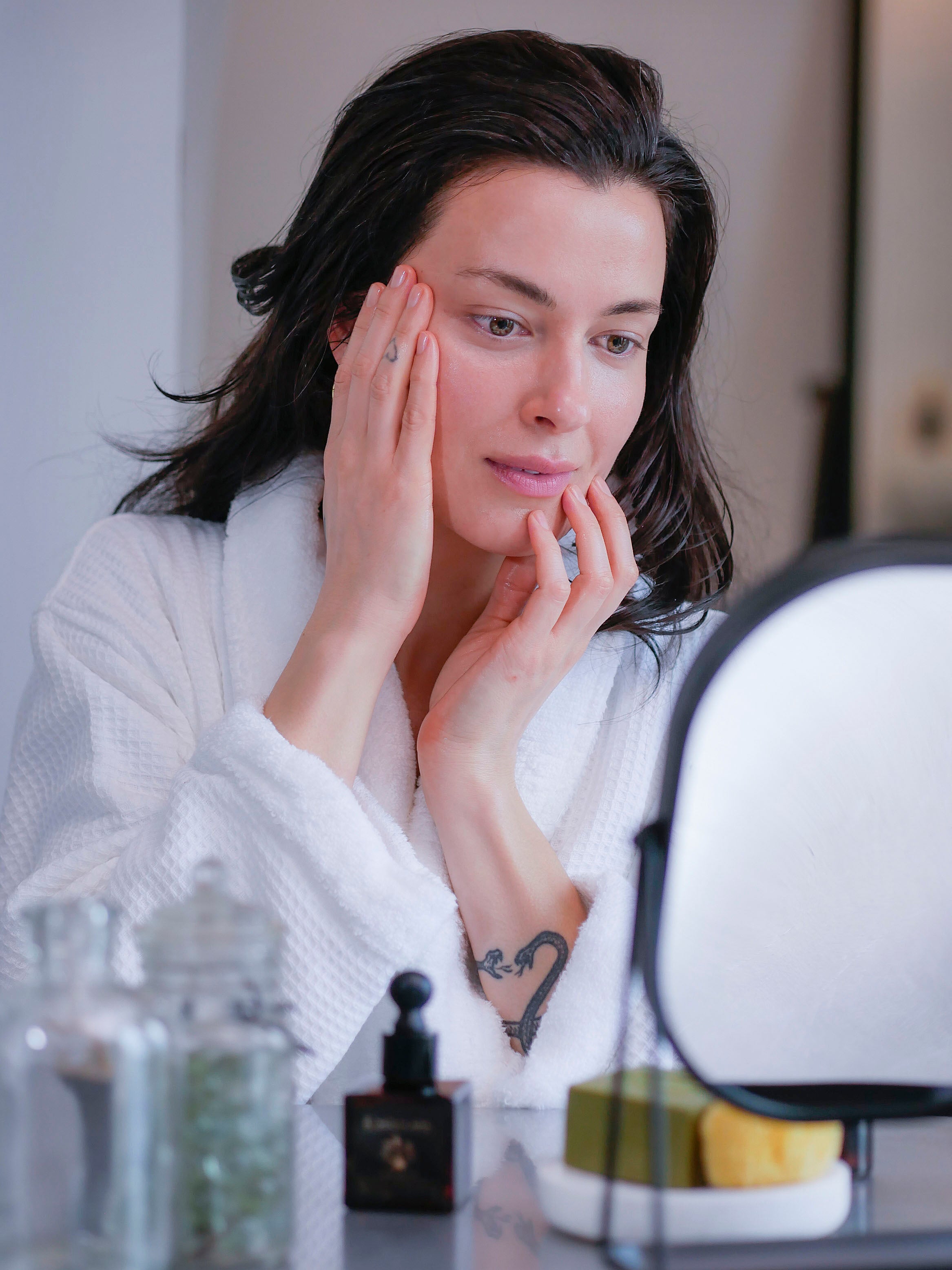 A woman with brown hair in a white fluffy bathrobe applying face serum for sensitive skin.