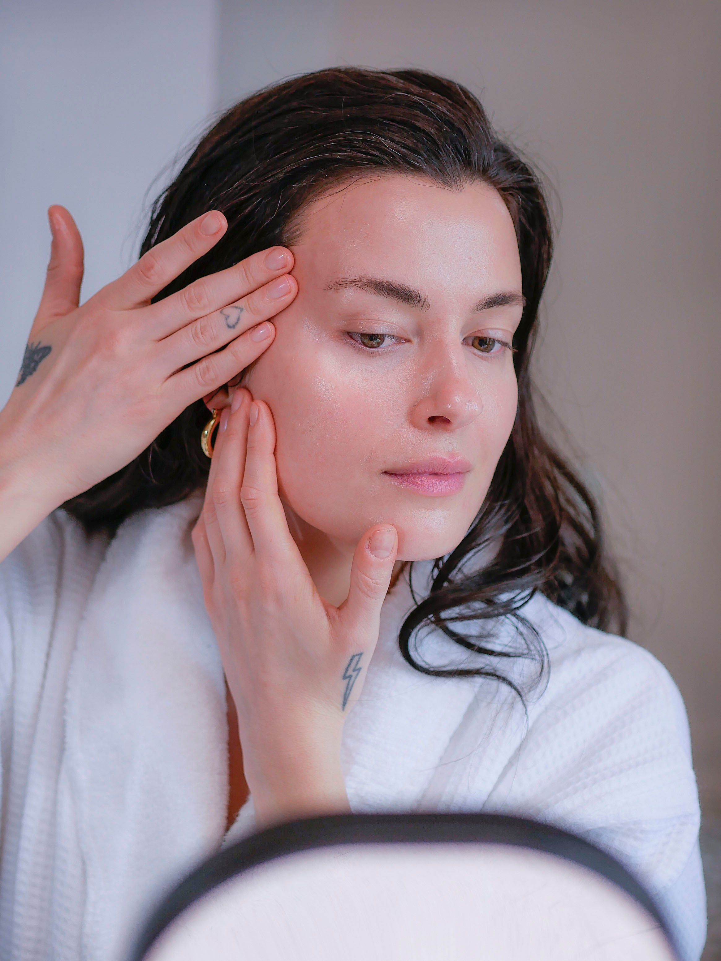 Woman with brown hair in a fluffy white bathrobe applying a face serum for combination skin.