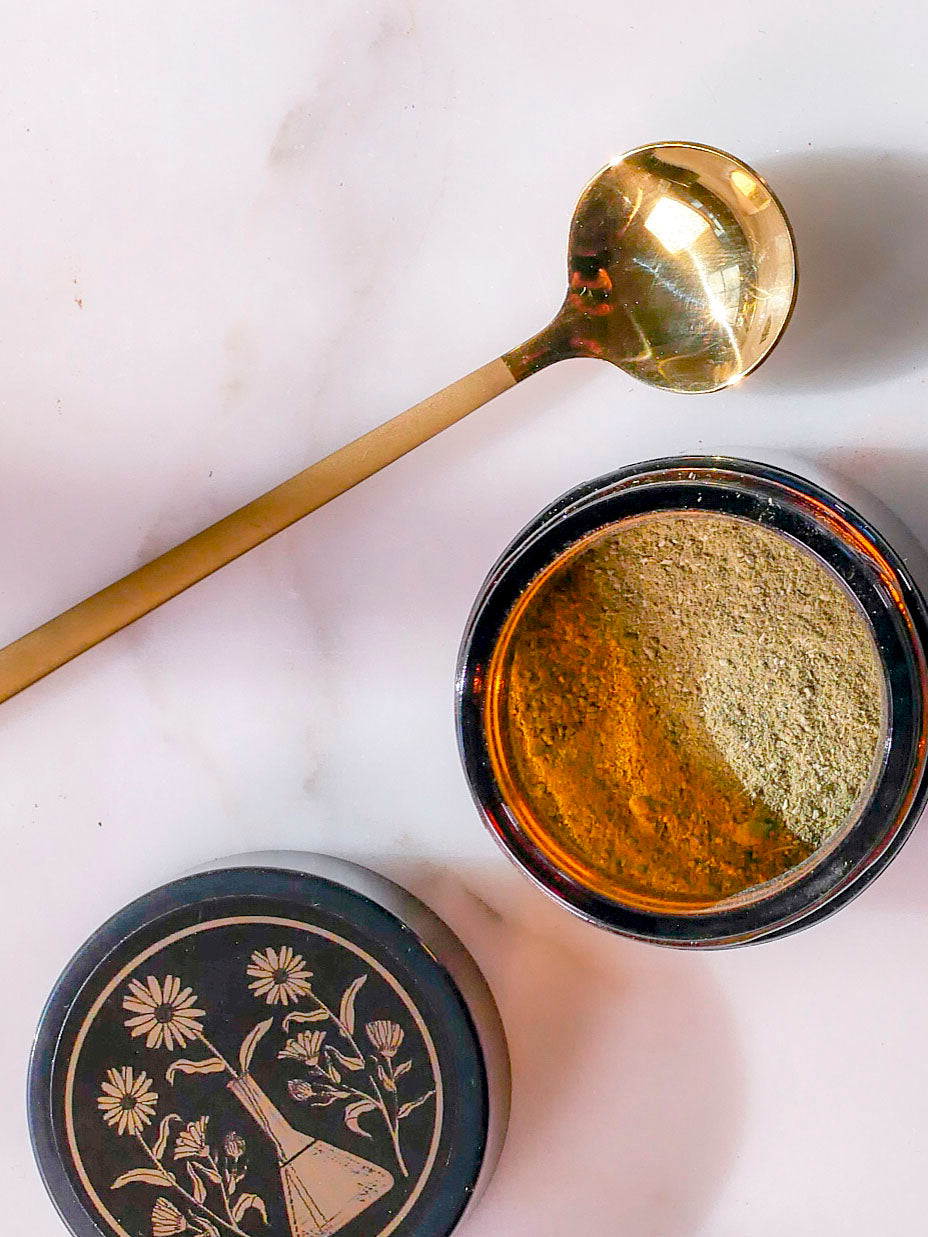 Gold face mask spoon on a marble counter next to a clay face mask container.